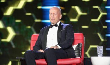 Bruce Willis îmbrăcat la sacou, poartă un papion negru și stă pe un scaun de culoare roșie, din piele