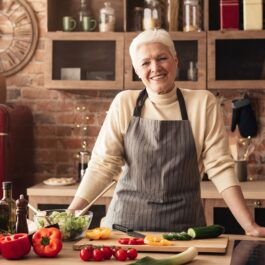 O femeie în vârstă se află într-o bucătărie bine utilată, poartă un șorț de bucătărie de culoare gri și se sprijină de un un blat de lucru