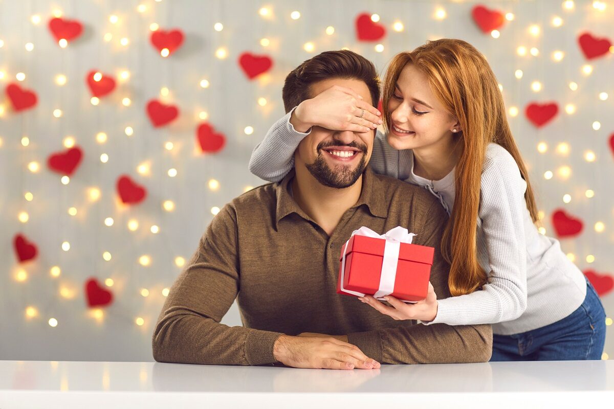Un cuplu fericit, format dintr-un bărbat care stă la o masă și o femeie care îi pune mâna la ochi și îi oferă un cadou de Ziua Îndrăgostiților