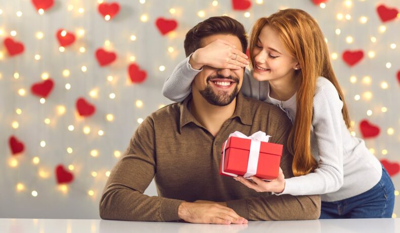 Un cuplu fericit, format dintr-un bărbat care stă la o masă și o femeie care îi pune mâna la ochi și îi oferă un cadou de Ziua Îndrăgostiților
