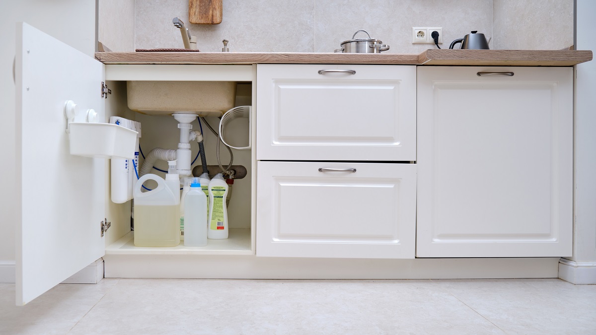 Un dulap de bucătărie, de culoare albă are ușa deschisă, văzându-se diferite țevi și obiecte depozitate sub chiuvetă