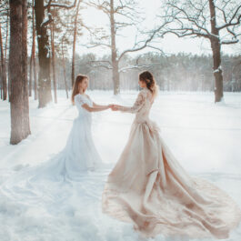 Două fete frumoase îmbrăcate în rochii albe lungi stau în zăpadă și se țin de mână