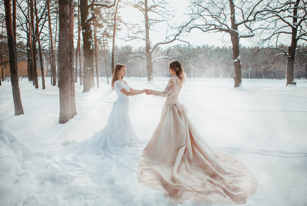 Două fete frumoase îmbrăcate în rochii albe lungi stau în zăpadă și se țin de mână