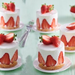 Cinci mini cheesecake cu căpșuni alături de un bol cu căpșuni și două furculițe