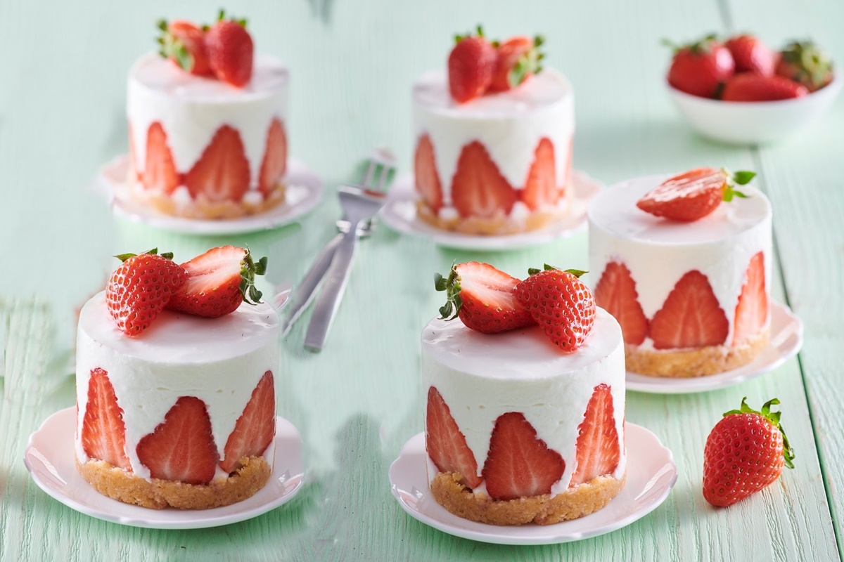 Cinci mini cheesecake cu căpșuni alături de un bol cu căpșuni și două furculițe