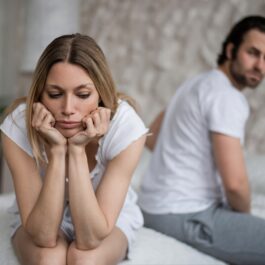 Un cuplu tânăr se află într-un dormitor, stând spate în spate, dezamăgiți de problemele din relația lor