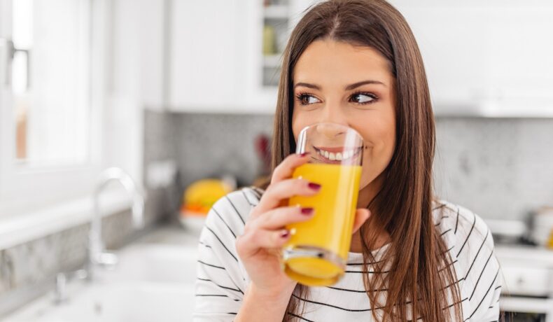 O femeie tânără care zâmbește consumă un pahar de suc de portocale natural