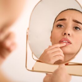 O femeie tânără și frumoasă stă mirată în fața unei oglinzi, îngrijorată de buzele sale crăpate