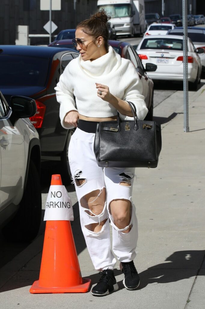 Jennifer Lopez este îmbrăcată lejer și poartă o pereche de blugi rupți de culoare crem, adidași de culoare neagră și părul prins în coadă
