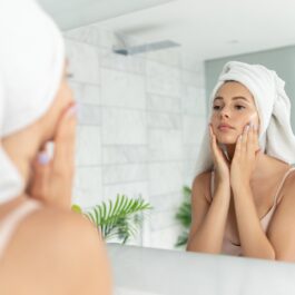 O femeie tânără și frumoasă poartă un prosop în cap și își masează încet pielea feței, privind în oglinda din baie