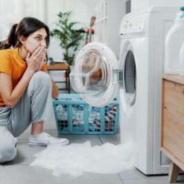 O femeie îngrijorată verifică de ce mașina ei de spălat nu mai funcționează și scoate multă spumă în exterior