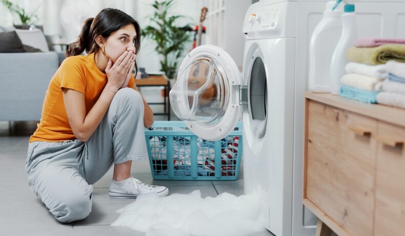 O femeie îngrijorată verifică de ce mașina ei de spălat nu mai funcționează și scoate multă spumă în exterior