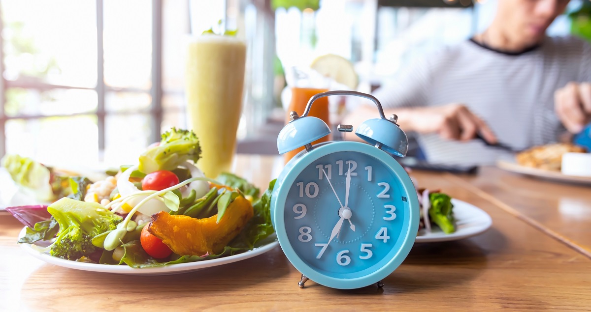 Un ceas deșteptător de culoare albastră, alături de o farfurie cu salată verde și carne slabă, pe o masă de lemn