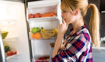 O femeie tânără și blondă stă gânditoare îîn fața unui frigider pentru a stabili rutina de dimineață