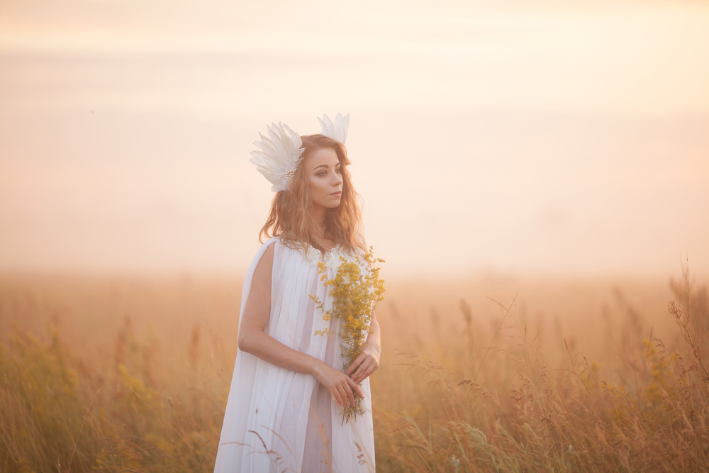 fată frumoasă îmbrăcată într-o rochie lungă, albă stă pe câmp cu un buchet de flori în mână