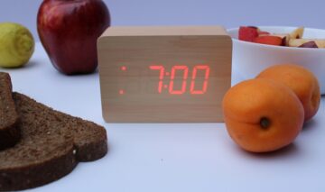 Un ceas care indică ora 7:00 dimineața, pus pe o masă cu fructe și pâine neagră