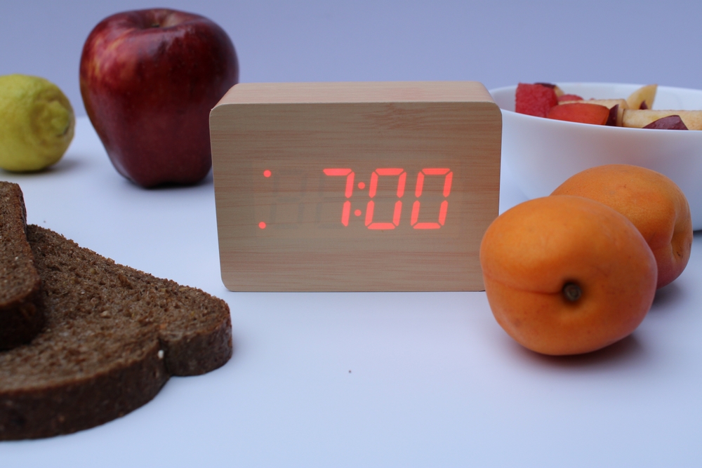 Un ceas care indică ora 7:00 dimineața, pus pe o masă cu fructe și pâine neagră