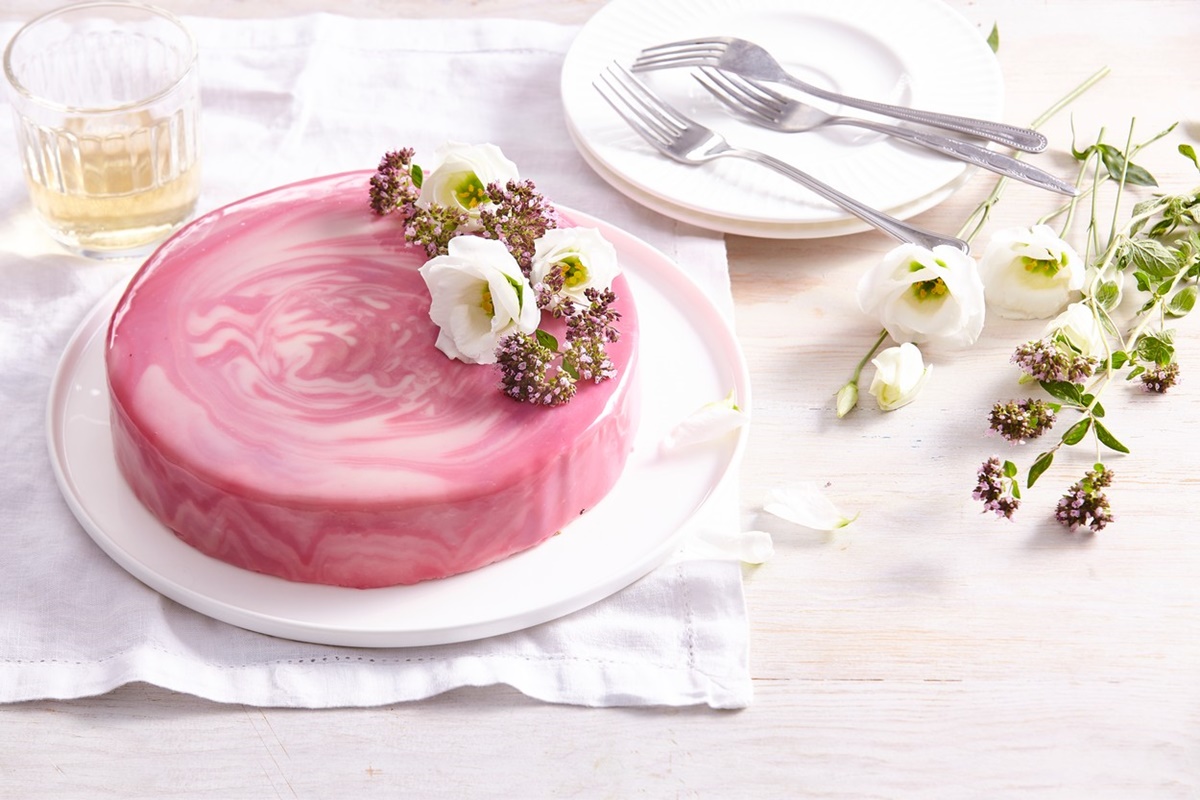 Tort de căpșuni cu glazură oglindă și flori comestibile pe un platou alb, alături de o farfurie cu tacâmuri
