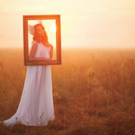 Fată frumoasă îmbrăcată într-o rochie lungă, albă stă pe un câmp și are o ramă în fața feței