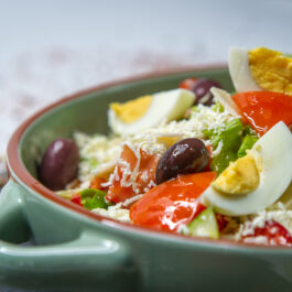 O salată grecească cu multe legume, ouă fierte și brânză rasă, într-un bol verde din ceramică
