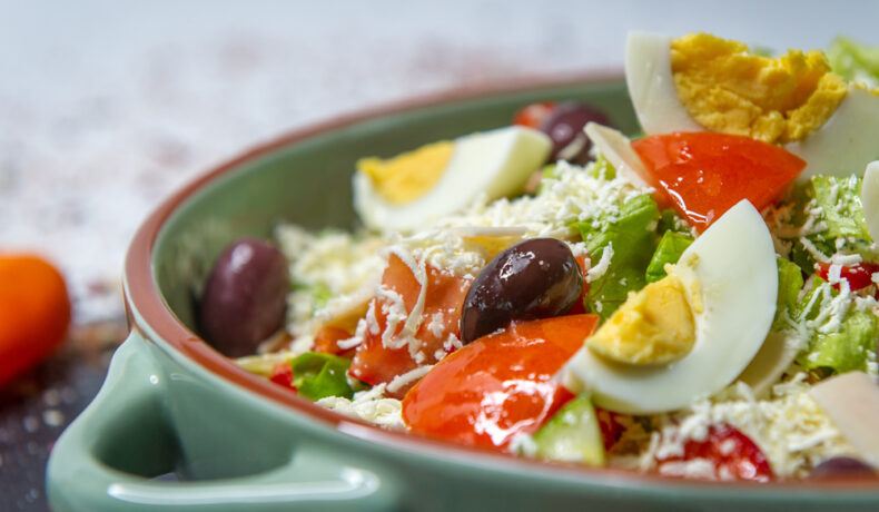 O salată grecească cu multe legume, ouă fierte și brânză rasă, într-un bol verde din ceramică