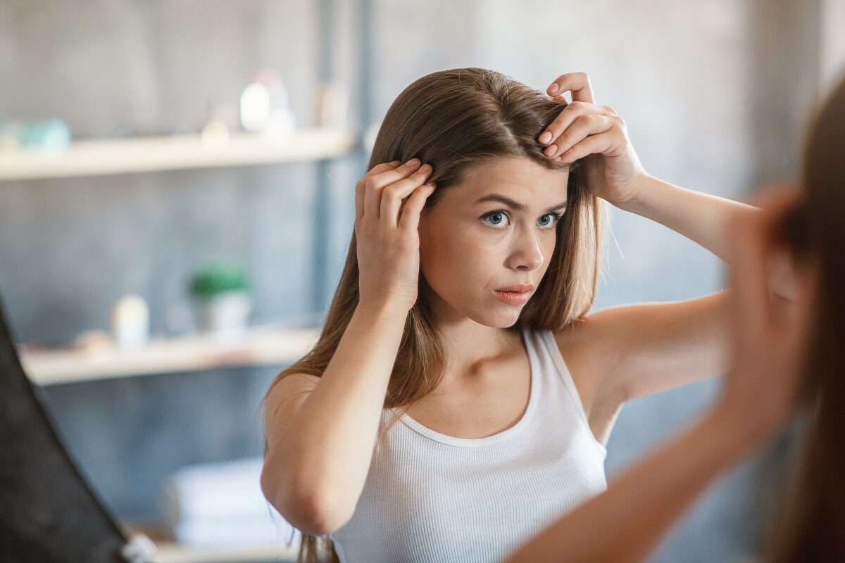 O tânăără se confruntă cu probleme serioase la nivelul scalpului în timp ce stă în fața unei oglinzi mari, verificându-și părul