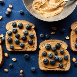 Un blat de bucătărie albastru pe care se află mai multe felii de pâine cu unt de arahide și afine pentru a ilustra una dintre principalele gustări care te pot ajuta să pierzi în greutate