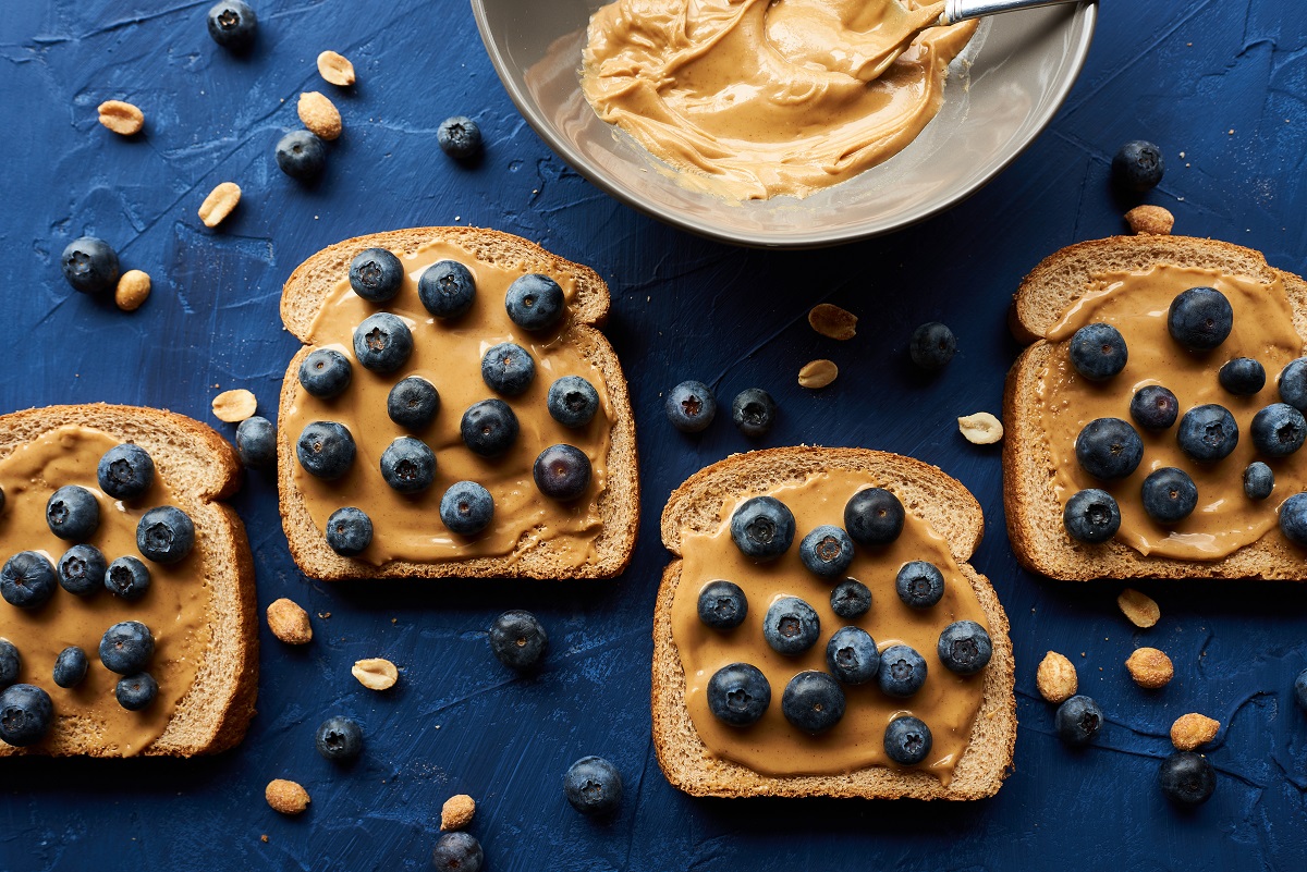 Un blat de bucătărie albastru pe care se află mai multe felii de pâine cu unt de arahide și afine pentru a ilustra una dintre principalele gustări care te pot ajuta să pierzi în greutate