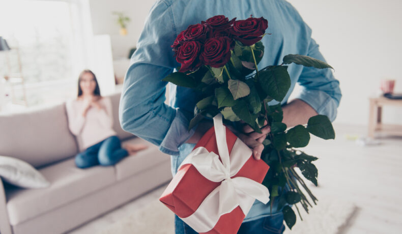 Bărbat cu flori în mână și cadou se îndreaptă spre partenera care stă pe canapea