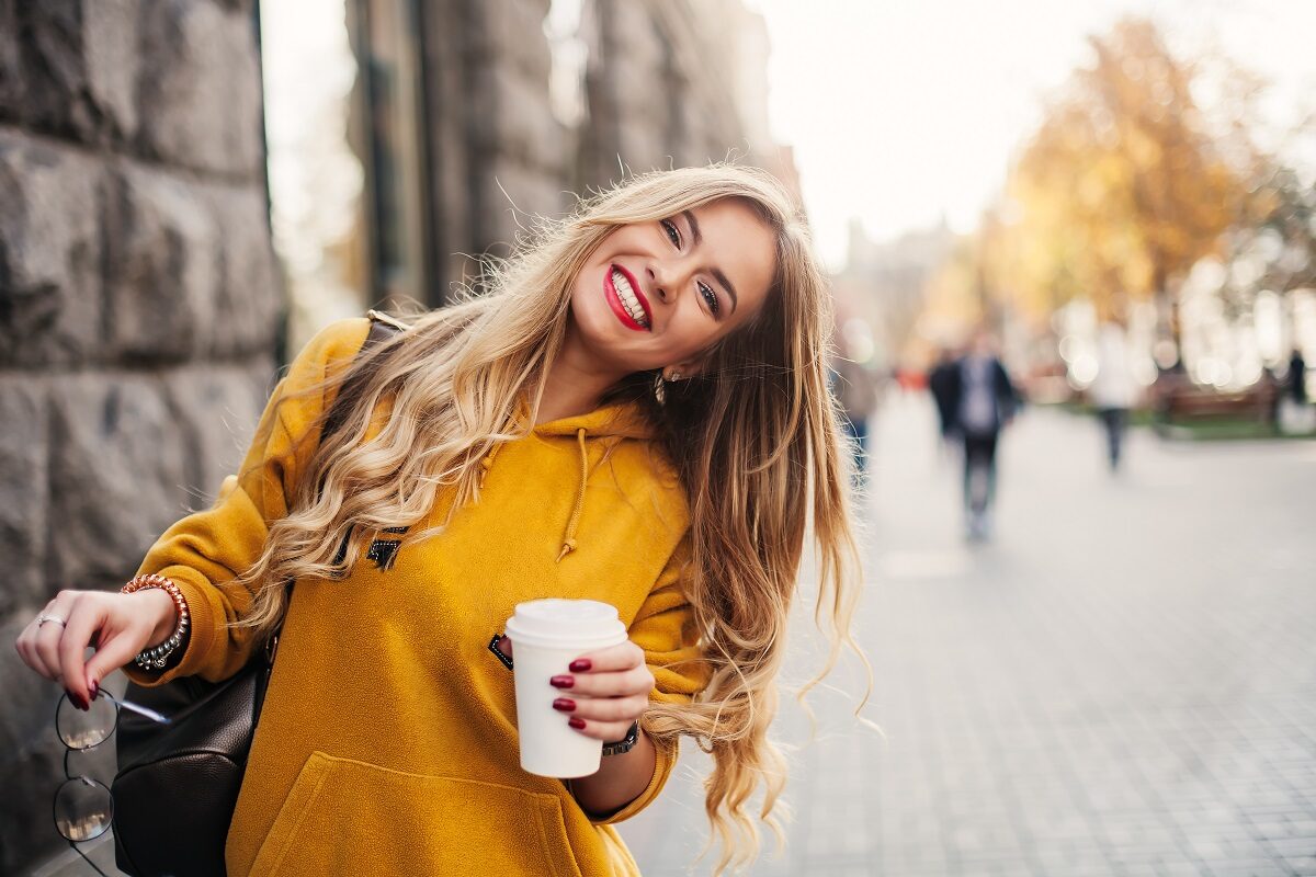 O femeie frumoasă, blondă, care zâmbește și ține în mână un pahar cu cafea