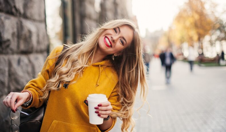 O femeie frumoasă, blondă, care zâmbește și ține în mână un pahar cu cafea