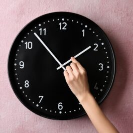 Un ceas negru pe un perete roz în timp ce este schimbat la ora de vară 2023 de o mână de femeie