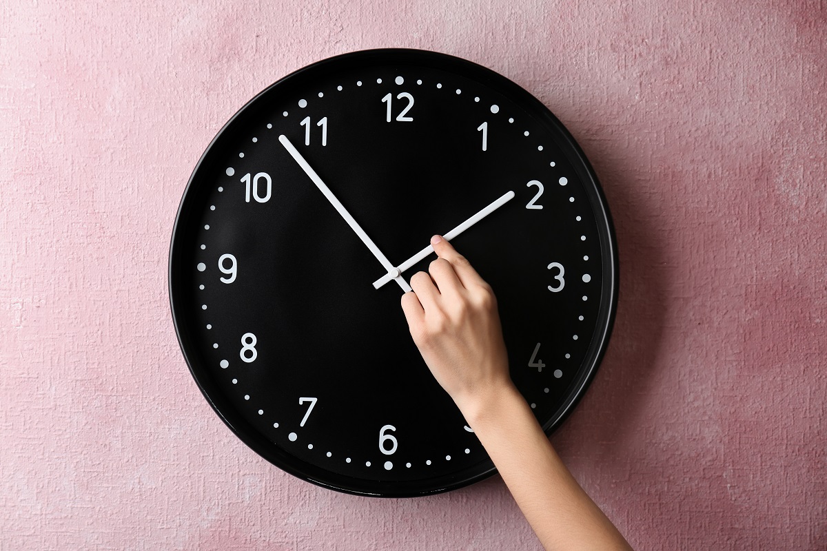 Un ceas negru pe un perete roz în timp ce este schimbat la ora de vară 2023 de o mână de femeie