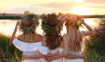 Tre fete îmbrăcate în rochii albe cu coronițe de flori pe cap privesc spre soare