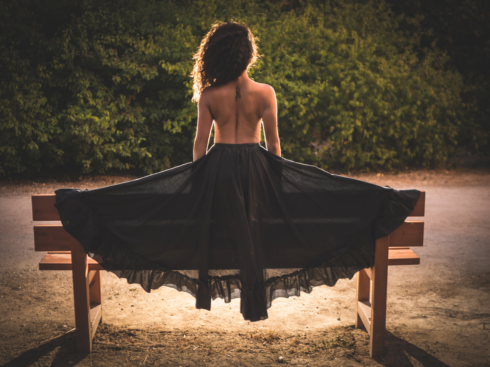 O femeie îmbrăcată în rochie neagră cu spatele gol, care stă pe o bancă, singură