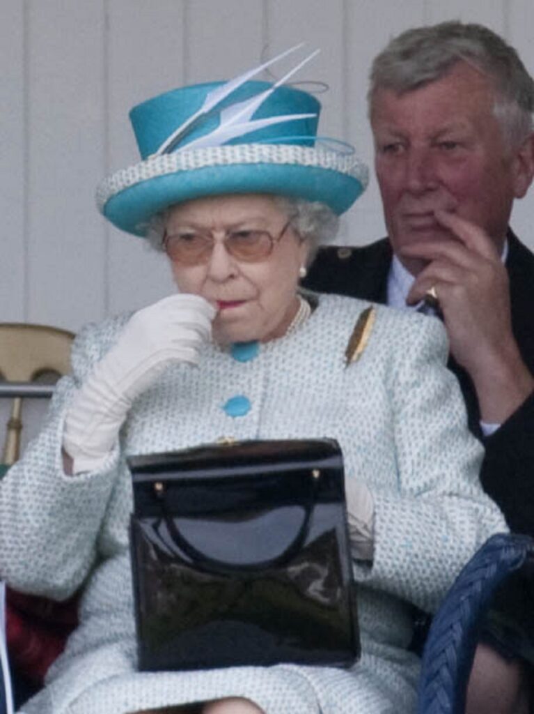 Regina Elisabeta ântr-un costum albastru în timp ce stă pe scaun și se dă cu ruj