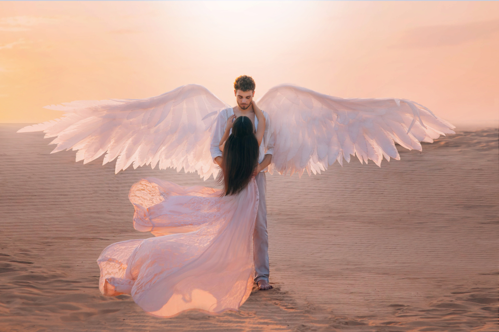 O femeie îmbrăcată într-o rochie diafană, care stă în brațele unui bărbat cu aripi de înger
