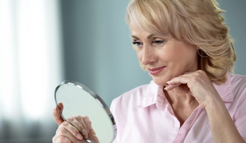 O femeie frumoasă, care are peste 50 de ani, ce se privește cu atenție într-o oglindă pe care o ține în mână