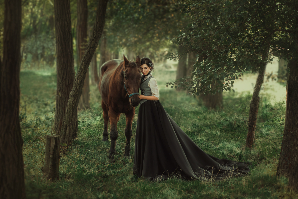 Fată frumoasă îmbrăcată într-o rochie lungă stă lângă un cal într-o pădure