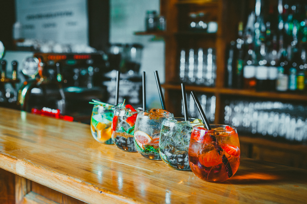 Cinci pahare cu câte un cocktail diferit, aranjate pe o tejghea lustruită într-un bar, plin de rafturi cu băuturi