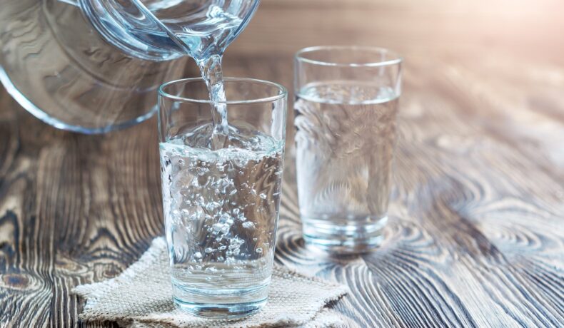 Două pahare de apă, pe o masă de lemn, în care se toarnă dintr-o carafă plină pentru a ilustra câte pahare de apă sunt recomandate zilnic