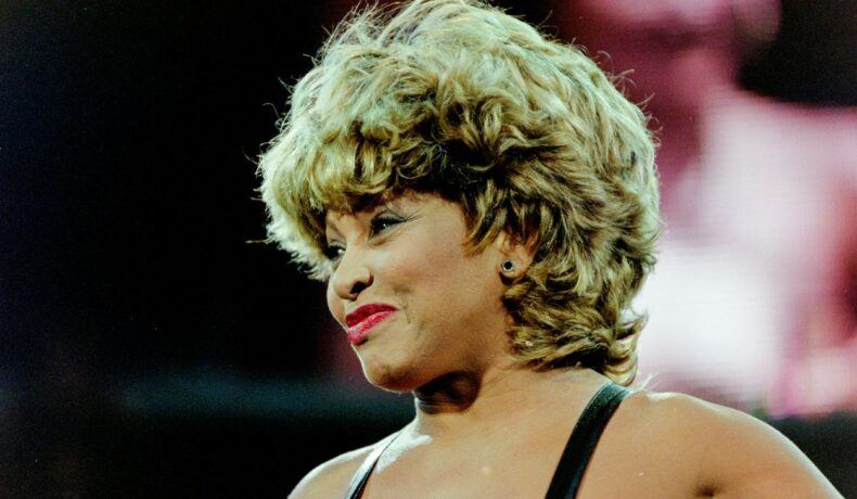 Tina Turner, într-o rochie din latex, neagră, cu bretele, pe scenă