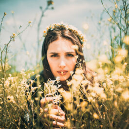 Fată frumoasă cu părul lung stă printre flori cu o coroniță de flori pe cap
