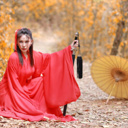 Fată frumoasă îmbrăcată într-un costum roșu stă pe jos într-o pădure cu o sabie în mână