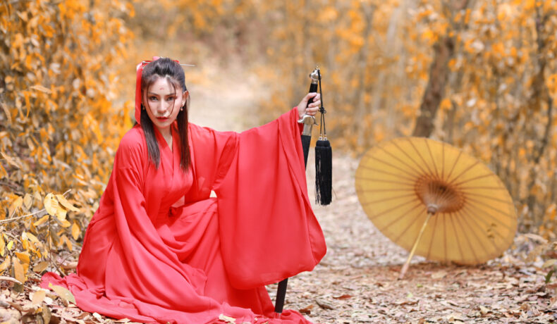 Fată frumoasă îmbrăcată într-un costum roșu stă pe jos într-o pădure cu o sabie în mână