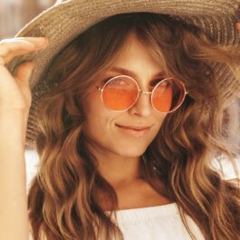 O femeie frumoasă cu ochelari de soare și pălărie care zâmbește la cameră pentru a ilustra una dintre principalele coafuri în trend vara aceasta