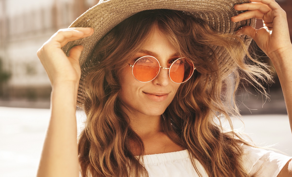 O femeie frumoasă cu ochelari de soare și pălărie care zâmbește la cameră pentru a ilustra una dintre principalele coafuri în trend vara aceasta