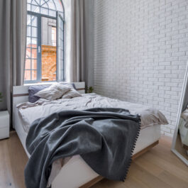 Un dormitor cu pat matriomonial, lejerie gri, ferestre mari, cu o oglindă mobilă în fața patului