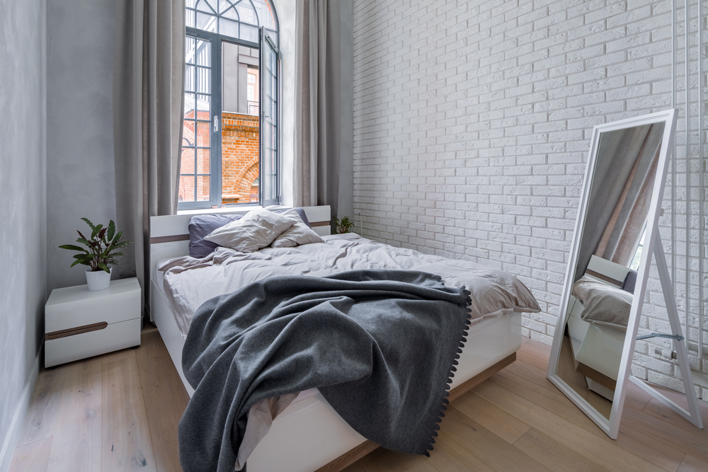 Un dormitor cu pat matriomonial, lejerie gri, ferestre mari, cu o oglindă mobilă în fața patului