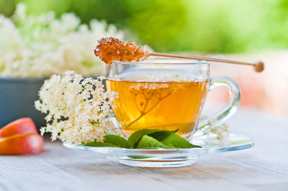 O cană transparentă cu ceai, cu o lingură de miere deasupra, pusă pe o farfurie transparentă, cu flori albe și frunze, toate așezate pe o masă de lemn